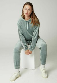 Set di loungewear in velluto a due pezzi di colore verde chiaro, composto da una felpa con cappuccio e pantaloni tapered; presenta strisce laterali argentate e polsini elasticizzati.