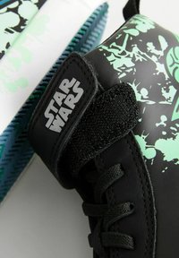 Schwarze Sneakers mit grünen Spritzmustern und einer glatten Textur. Merkmale sind ein Klettverschluss und Schnürsenkel, mit einem "Star Wars"-Logo an der Seite.