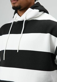 Sweat à capuche rayé noir et blanc avec cordon de serrage, doté d'une texture douce en coton et d'une coupe décontractée. Rayures horizontales prononcées sur tout le tissu.