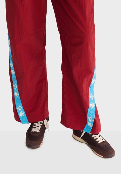 Pantalon rouge à jambes larges avec des bandes bleues ornées de motifs blancs de style pixel, associé à des baskets marron foncé à lacets blancs sur un fond clair.