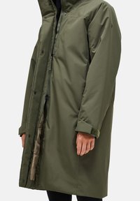 Olijfgroene, knielange, gewatteerde jas met een hoge kraag, voorrits en drukknoopjes. Beschikt over fleece-voering bij de manchetten en een binnenzak.