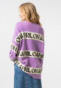 Gros pull en tricot violet surdimensionné avec des bandes blanches contrastantes comportant du texte. Texture douce et coupe décontractée. Assorti à une jupe en denim.
