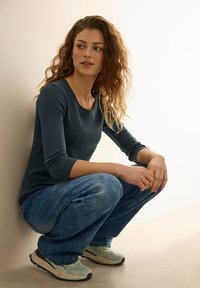 Dlouhý rukáv tmavě modré tričko, světle modré vybledlé džíny a béžové tenisky s černými podrážkami. Subjekt se schrbuje proti zdi.