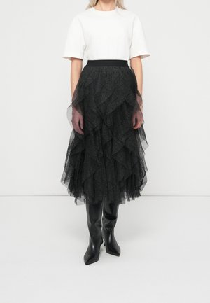 Personne portant un haut blanc à manches courtes, une jupe midi en tulle noire superposée et des bottes noires pointues montant jusqu'aux genoux, debout devant un fond uni.