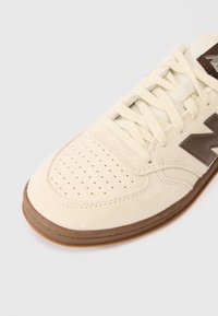 Kremowe zamszowe i skórzane sneakersy z perforowanym noskiem, płaskimi sznurówkami i brązową gumową podeszwą. Posiadają kontrastowe brązowe logo na boku.