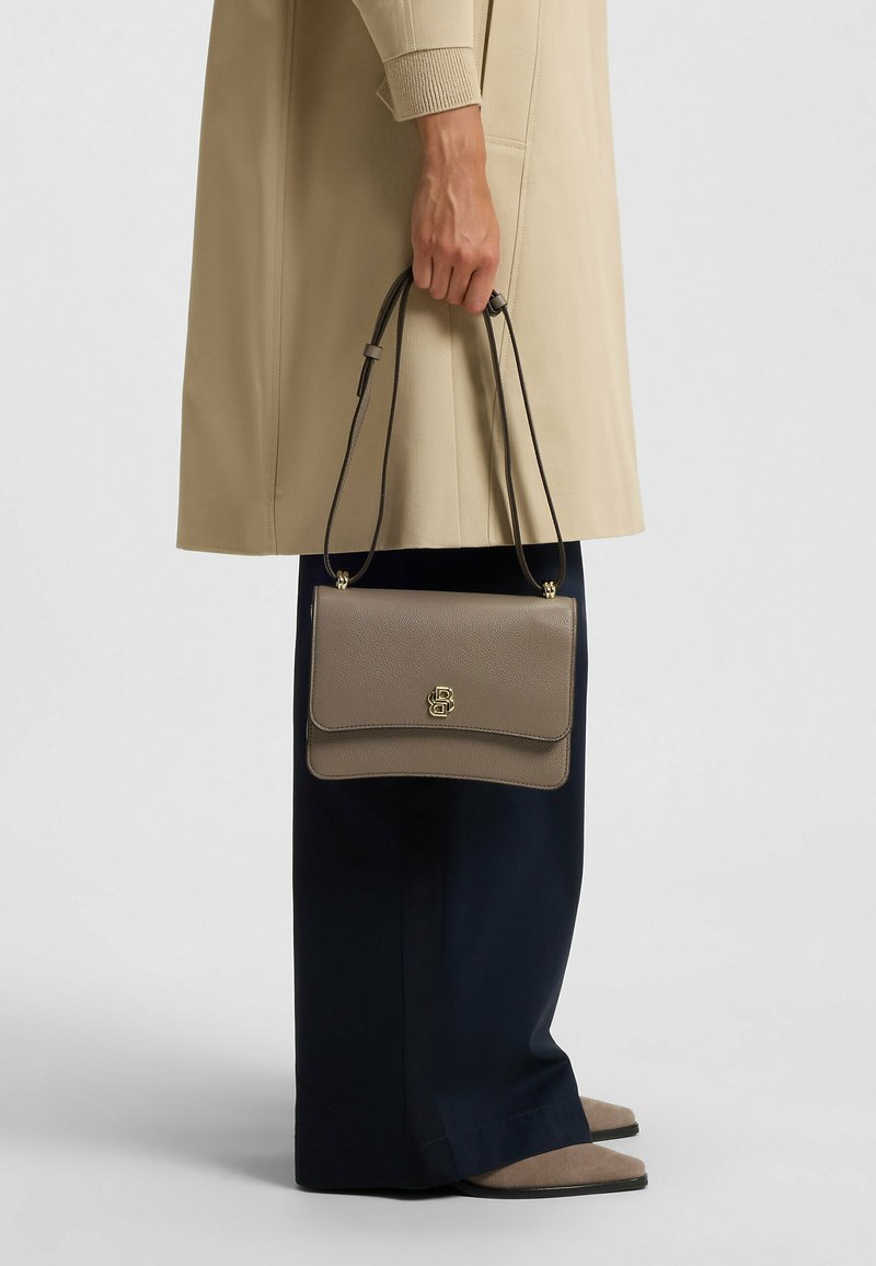 Borsa a tracolla in pelle testurizzata grigia con chiusura a logo dorato, design semplice e tracolla regolabile, tenuta da una persona con un cappotto beige.