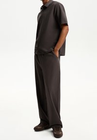 Mörkgrå kortärmad skjorta med knappar i texturerad tyg, tillsammans med löst sittande mörkgrå byxor och bruna träningsskor.