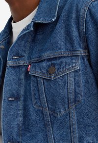 Veste en denim bleu foncé avec des motifs ton sur ton complexes, col classique, quincaillerie en bronze et poches latérales. Détails de couture remarquables.