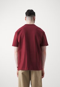 Burgundské bavlněné tričko s krátkým rukávem a kulatým výstřihem; jednoduchý design s volným střihem, zobrazené ze zadní strany na bílém pozadí.