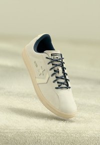 Beige semsket low-top sneakers med marineblå snørebånd og merke. Har en stjerne-logo og en gummisåle. Glatt tekstur og strømlinjeformet design.