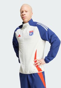 Chaqueta deportiva blanca y azul con acentos rojos, que presenta un cierre de cremallera y el logo del Olympique Lyonnais en el pecho. Tejido texturizado, mangas largas.