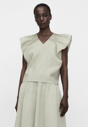 Kobieta ubrana w jasnobeżową sukienkę bez rękawów z falbanianymi ramionami i plisowanymi detalami na górze i spódnicy, na jednolitym tle.