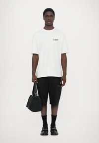 T-shirt en coton blanc avec un petit logo noir, shorts noirs, sac noir avec une bandoulière, chaussettes noires et chaussures noires avec un accent métallique.