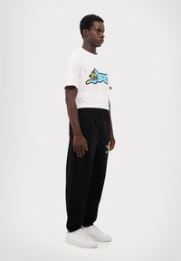 White t-shirt with a blue cartoon cat print; black sweatpants with an embroidered yellow sun icon; white sneakers; minimal athletic style.