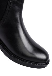 Primo piano della punta in pelle nera liscia e lucida e della suola cucita di un Chelsea boot su uno sfondo bianco.