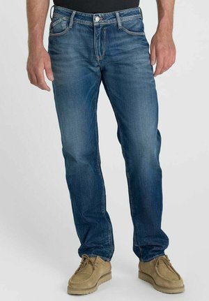 Uomo che indossa jeans blu a gamba dritta e scarpe casual beige, in piedi davanti a uno sfondo chiaro e uniforme.