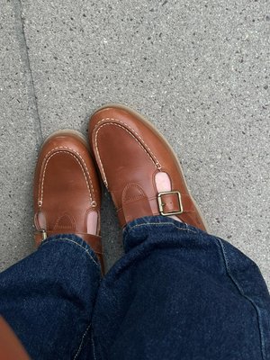 Καφέ δερμάτινα παπούτσια με αγκράφες φορεμένα με μπλε τζιν, που στέκονται σε γκρι τσιμεντένιο πάτωμα.