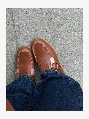 Καφέ δερμάτινα παπούτσια με αγκράφες φορεμένα με μπλε τζιν, που στέκονται σε γκρι τσιμεντένιο πάτωμα.