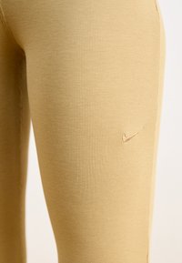 Beige ribbota bikses izgatavotas no elastīga auduma, ar mazu, iegravētu Nike logo netālu no gurniem. Gluda tekstūra ar pieguļošu dizainu.