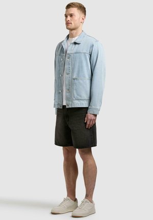 Mann iført lyseblå jeansjakke, hvit t-skjorte, svarte jeansshorts og kremfargede joggesko, står mot en ensfarget bakgrunn.