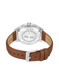 Reloj Timberland con caja de acero inoxidable, correa de cuero marrón y hebilla plateada. La parte posterior presenta un logo de árbol y detalles de resistencia al agua.