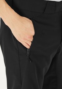 Zwarte broek met een ritssluiting aan de zijkant. Het materiaal lijkt lichtgewicht en soepel. Het ontwerp is getailleerd, met een reliëf logo-detail.