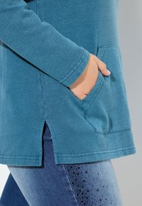 Mano infilata nella tasca anteriore di una maglietta teal a maniche lunghe con spacchi laterali, abbinata a jeans blu con decorazioni in perline nere.