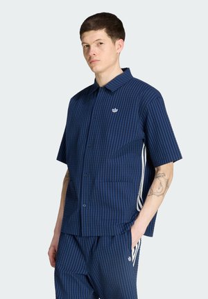 Joven con cabello corto y oscuro lleva una camisa de botones de manga corta azul a rayas y pantalones a juego con franjas blancas en los costados, de pie con la mano en el bolsillo.