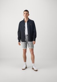 Camisa de mezclilla abotonada en azul oscuro, combinada con una camiseta blanca sin mangas, pantalones cortos enrollados gris claro y zapatillas blancas con suelas marrones.