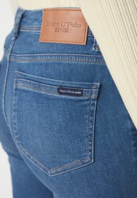 Blugi din denim albastru cu o etichetă din piele maro inscripționată "Marc O'Polo DENIM." Prezintă cusături aurii și un design minimalist pentru buzunarul din spate.