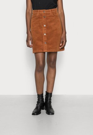 Femme portant une jupe en velours côtelé marron boutonnée, un haut noir et des bottines noires, debout devant un fond clair uni.