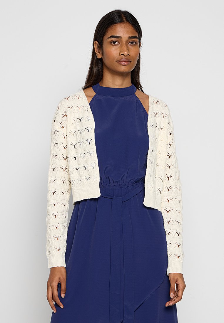 Kremowy, koronkowy kardigan z otwartymi wzorami i długimi rękawami, noszony na ciemnoniebieską sukienkę z wcięciem w talii i wysokim dekoltem.