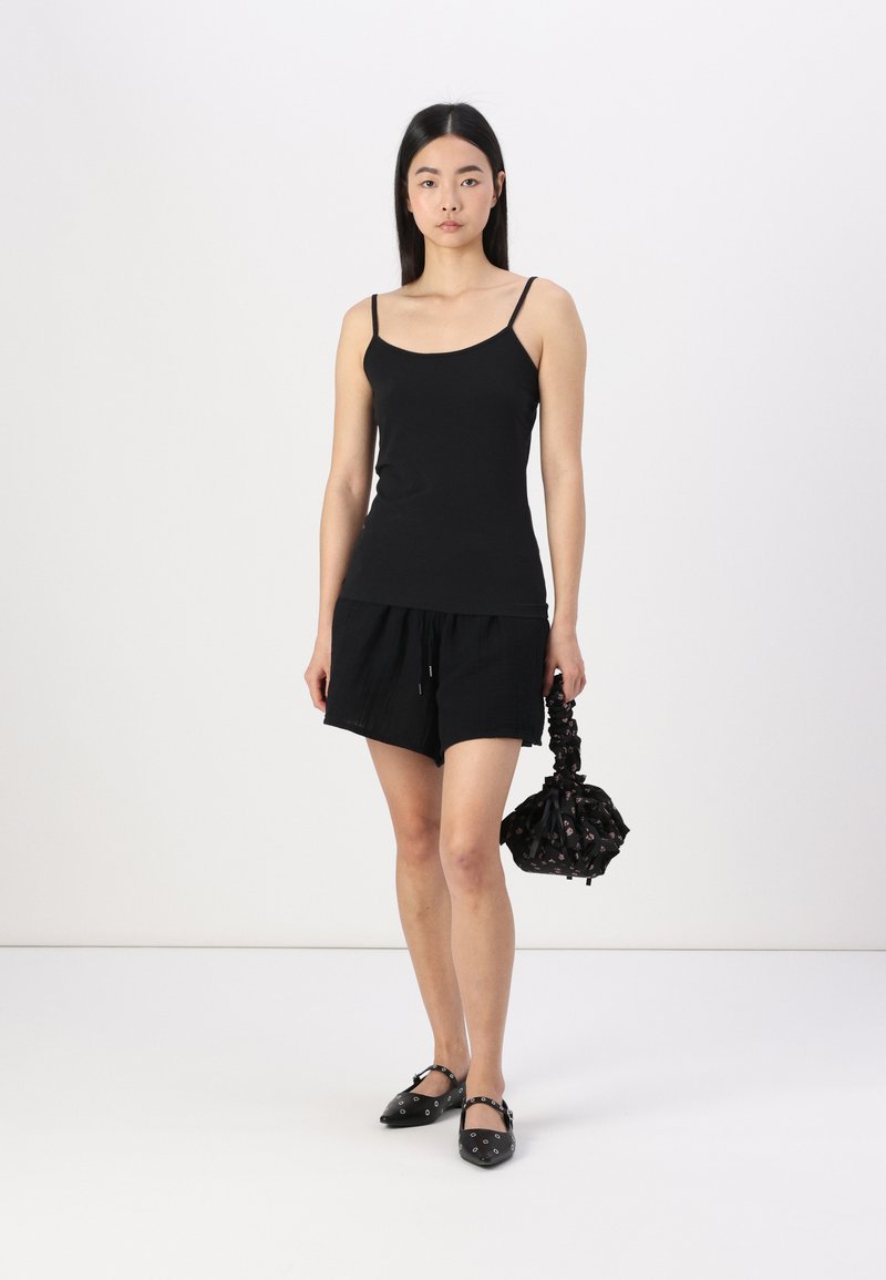 Black sleeveless top, paired with black shorts. Model holds a black patterned bag, wearing black flats with hardware accents. Simple, cohesive look.