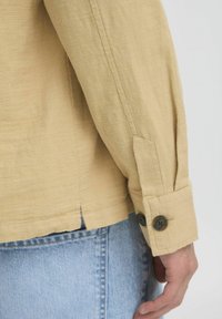 Chemise en lin beige à manches longues, avec un poignet comportant deux boutons foncés et une petite fente sur le côté à l'ourlet. Portée sur un jean bleu clair.