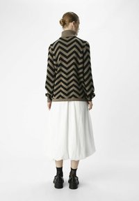 Maglione a zigzag marrone e nero con colletto alto, indossato su una gonna bianca, abbinato a scarpe nere e calzini alla caviglia.