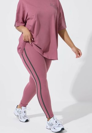 Person iført lilla leggings med sorte sidelommer, matchende løstsiddende t-shirt og hvide sneakers, der går mod en ensfarvet baggrund.