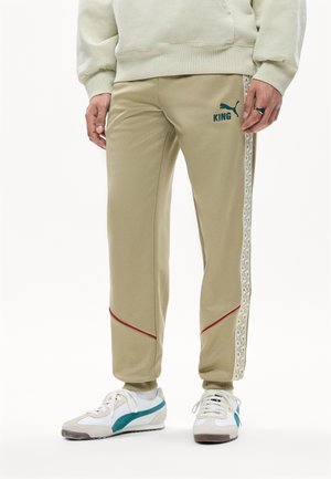 Beige Puma træningsbukser med mønstrede sidestriber, rød piping nær knæene, båret med hvide sneakers med turkis accenter.