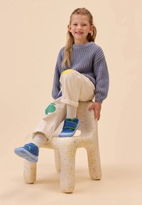 Blaue und grüne Kinderschuhe mit kuscheliger Textur; cremefarbene Hose mit bunten Obstmotiven; gestrickter grauer Pullover; sitzt auf einem gepunkteten Stuhl.