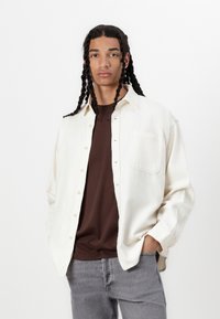 Camicia oversize color crema con taschino, chiusura a bottoni, indossata su una t-shirt marrone, abbinata a jeans grigi.