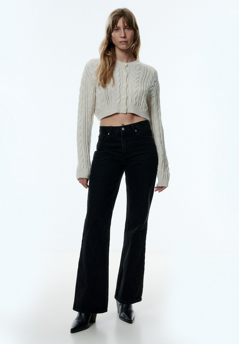 Cardigan en tricot torsadé crème raccourci avec des boutons, associé à un jean noir évasé taille haute et des bottines à talons noires.
