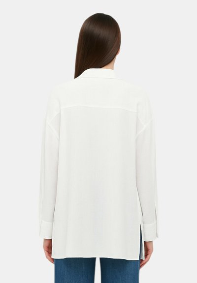 Camicia bianca a maniche lunghe con colletto, vestibilità rilassata, spacchi laterali e tessuto testurizzato. La vista posteriore mostra linee morbide e dettagli sottili.