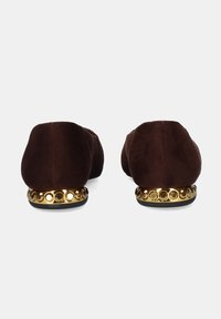 Zapatos planos de ante marrón con un acento circular dorado decorativo en el tacón, con punta redondeada y textura suave.