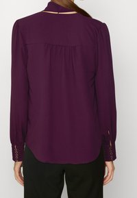 Kobieta w fioletowej bluzce z długim rękawem i mankietami zapinanymi na guziki, widoczna od tyłu, w połączeniu z czarnymi spodniami.