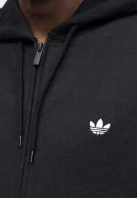 Gros plan sur une personne portant un sweat à capuche noir zippé avec des cordons noirs et un petit logo blanc Adidas trefoil sur la poitrine.