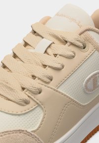 Μπεζ και λευκό αθλητικό παπούτσι με παχιά υφασμένα κορδόνια, λογότυπο Champion στο πλάι και μπεζ σουέντ κάλυμμα στα δάχτυλα, εμφανιζόμενο σε κοντινή λεπτομέρεια.