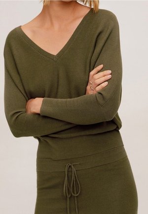 Femme portant un pull vert olive à col V manches longues et une jupe assortie avec une taille à cordon, bras croisés sur la poitrine.
