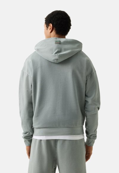 Mann trägt hellgrauen Kapuzenpullover und passende Jogginghose, von hinten vor schlicht weißem Hintergrund gesehen.