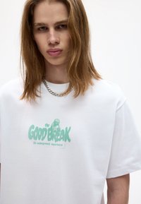 Valkoinen puuvillapaita, jossa on vihreä graafinen teksti "the GOOD BREAK" ja "the underground experience". Pyöreä kaula-aukko ja rento istuvuus.