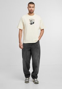 Cremefarvet bomulds t-shirt med grafisk tryk på brystet, sat sammen med løstsiddende mørke grå jeans og sorte sneakers med hvide detaljer.