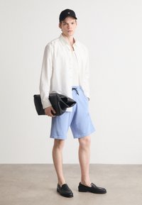 Lett hvit skjorte med knapper, kremfarget singlet, blå shorts med snor, svart caps, overdimensjonert svart veske, vevde svarte sko.